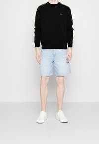 Svart sweatshirt med gula sidostråkar, rundad halsringning och logotyp. Ljust blå denimshorts med uppvikta fållar. Vita sneakers. Minimalistisk bakgrund.