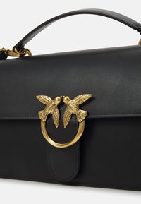 Čierna kožená kabelka s pevnou konštrukciou, hornou rukoväťou a zlatistým zapínaním s dvoma motívmi vtákov. Hladký povrch, minimálne prešívanie.