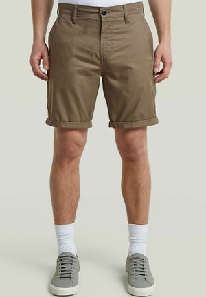 Homme portant un short marron retroussé, des chaussettes blanches et des baskets grises, debout devant un fond neutre.
