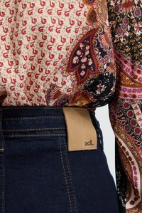 Blusa de tejidos estampados con diseños florales y de paisley en negro, rojo y beige, superpuesta sobre unos jeans de mezclilla oscuros con una etiqueta de cuero marrón.