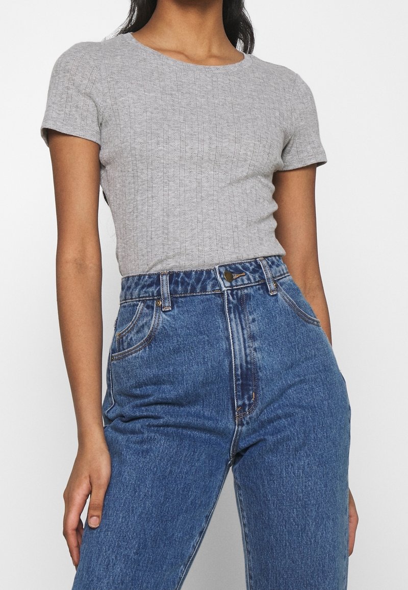 Femme portant un haut gris côtelé à manches courtes, rentré dans un jean en denim bleu taille haute, les mains partiellement dans les poches.