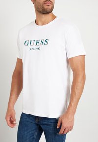 T-shirt en coton blanc avec un logo "GUESS" brodé en vert et un motif texturé. Encolure ronde classique et manches courtes.
