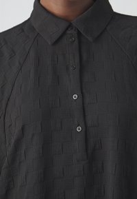 Chemise noire texturée avec col et patte de boutonnage sur le devant, partiellement boutonnée, portée par une personne à la peau foncée.