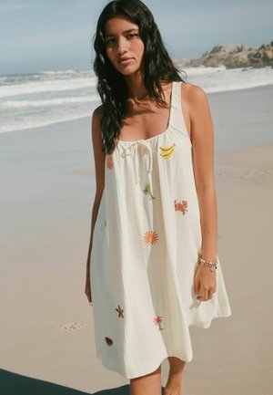 Young woman with long dark hair wearing a white sundress with colorful tropical embroidery, standing barefoot on a sandy beach.
