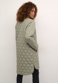 Lang quilted frakke i dæmpet grøn, med diamantformet syninger, sidelommer og en rundet kant. Blødt materiale med en glat overflade.
