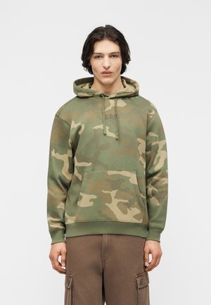 Joven con cabello oscuro de longitud media, vestido con una sudadera con capucha de camuflaje verde y marrón y pantalones cargo marrones, de pie frente a un fondo liso.