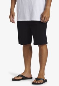 Black cotton shorts with a straight fit, knee-length, paired with black flip-flops. Model wears a white short-sleeve shirt.