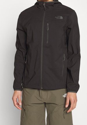 Osoba nosi czarną kurtkę z kapturem na zamek oraz oliwkowe spodnie, obie z logo The North Face na klatce piersiowej i udzie.