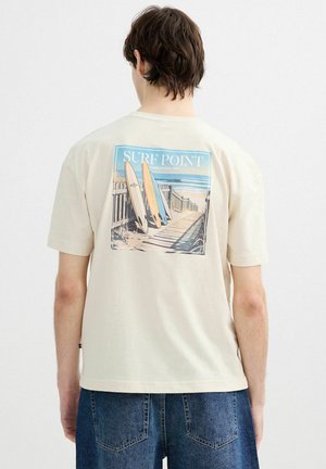 Persona que lleva una camiseta beige con un estampado de una escena de playa que muestra tres tablas de surf apoyadas contra una cerca y el océano al fondo.