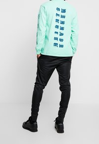 Mintsävyinen pitkähihainen paita, jossa on graafinen teksti selässä, yhdistettynä mustiin istuviin housuihin. Paita on rentoa mallia ja tyyliltään arkinen.