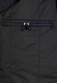 Sezione di giacca a vento nera con una fascia nera orizzontale che presenta un accento rosso, bianco e blu sotto un pulsante circolare nero.