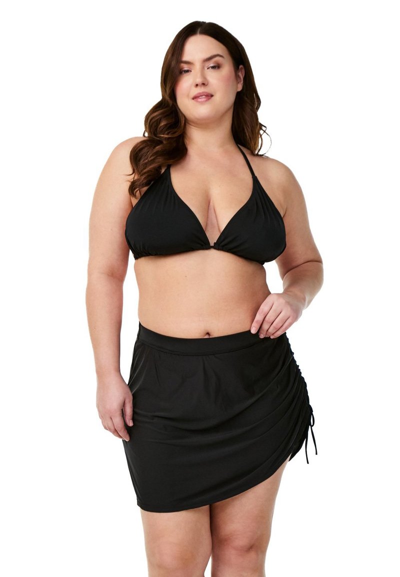 Plus izmēra sieviete pozē melnā haltera bikini augšpusē un burkšķī apakšā uz baltas fona.