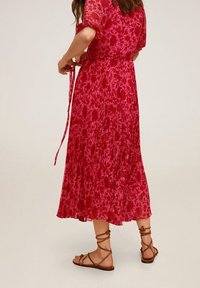 Robe florale rouge avec des manches courtes et une taille cintrée, présentant une jupe plissée et fluide. Portée avec des sandales à talons bruns.