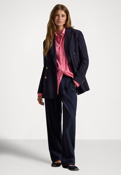 Sieviete stāv, ar gariem matiem, valkājot rozā kreklu ar pogām, tumši zilu divrindu žaketi, brīva piegriezuma bikses un melnas ērtas kurpes.