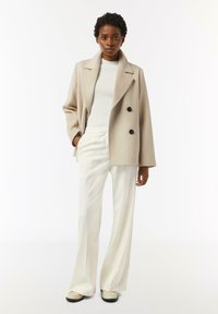 Beige double-breasted coat with black buttons, white ribbed sweater, and white wide-leg trousers; model stands with one hand in pocket.