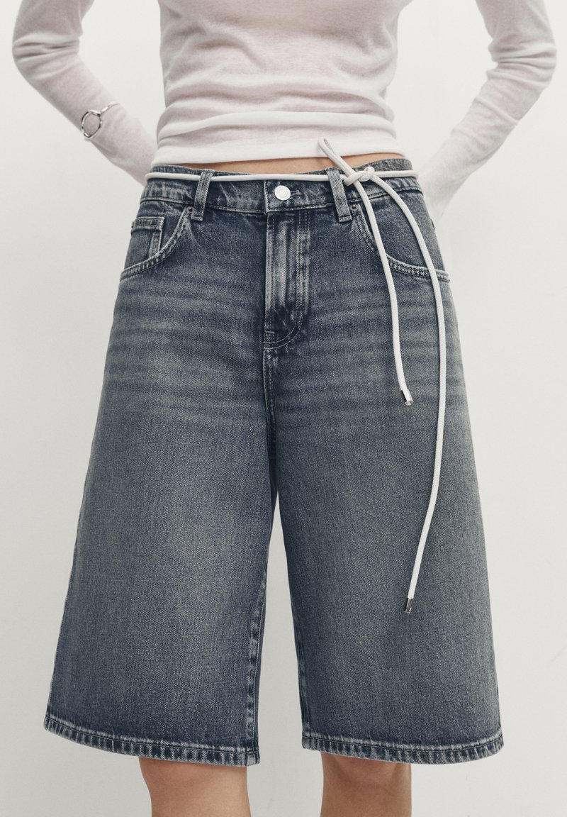 Personne portant un short en jean bleu foncé, taille haute, ample, avec une ceinture en corde blanche nouée à la taille, associé à un haut à manches longues clair.