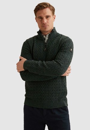 Hombre con cabello castaño claro que lleva un suéter de punto trenzado verde oscuro con cremallera y pantalones negros, brazos cruzados, expresión neutral.