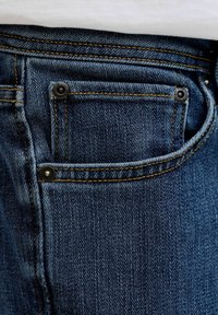 Tmavě modré džínové kalhoty zblízka zobrazující malou přední kapsu na mince s oranžovým prošíváním a černými kovovými nýty.