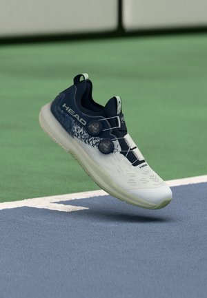 Hvit og marineblå atletisk sko med teksturert overdel, BOA-snøresystem og gummisåle, plassert mot en grønn og blå banebakgrunn.