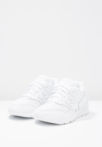 Bílé sportovní boty s perforovanými detaily, kulatou špičkou, texturovanou tkaninou a gumovou podrážkou; obsahují šněrování a značkování na jazyku a boku.