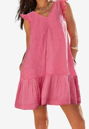 Kvinde iført ærmeløs pink kjole i krøllet stof med flæsekant og skulderdetaljer, accessoriseret med lagdelte halskæder og armbånd.