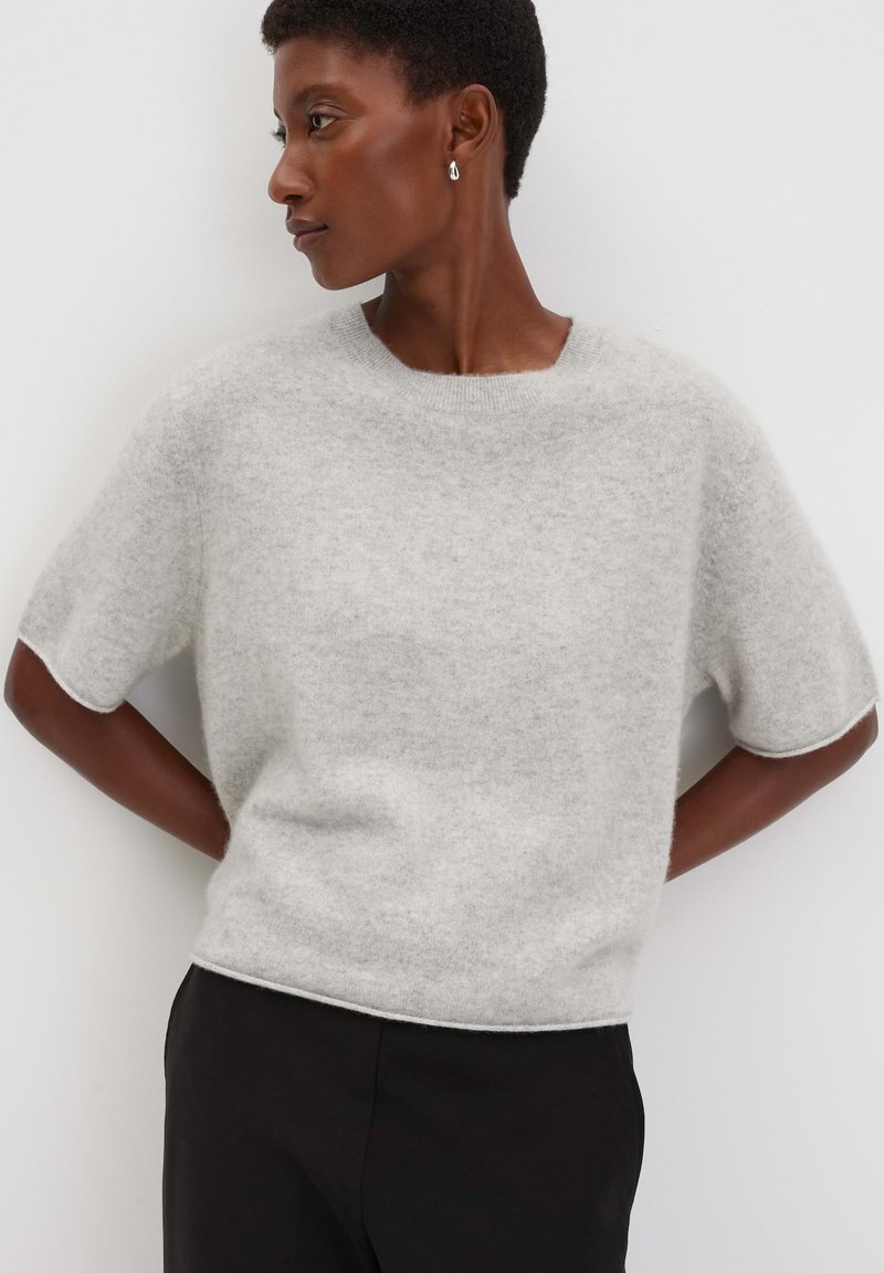 Vrouw met kort haar die zilveren hangende oorbellen draagt, een lichtgrijze trui met korte mouwen van pluizig materiaal en een zwarte broek, terwijl ze naar links kijkt.