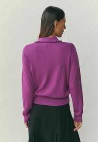 Purple knit sweater featuring a ruffled collar, balloon sleeves, and a fitted hem, worn over a black skirt. Smooth texture.