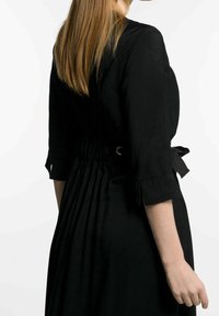 Svart klänning med figursydd midja med rynkor, trekvartslånga ärmar och en knytning bak. Slät, texturerad tyg.