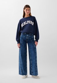 Marineblå sweatshirt med "SCALPERS" i hvid tekst, parret med højtaljede, vide blå denim jeans med en falmet finish og synlige sømme.