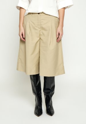 Lys beige culottes med vid ben, parret med skinnende sorte knæhøje støvler med spids tå, båret med en hvid langærmet skjorte.
