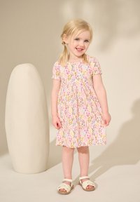 Robe courte à manches courtes florale rose avec des fleurs aux couleurs pastel et des manches à volants, associée à des sandales blanches. Le fond présente une teinte neutre claire.