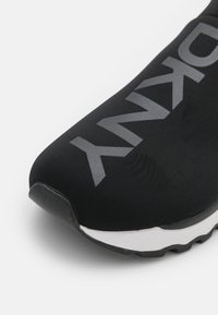 Gros plan sur la pointe d'une sneaker noire sans lacets avec un texte gris en gras sur la tige et une semelle intermédiaire blanche avec une semelle extérieure noire texturée.