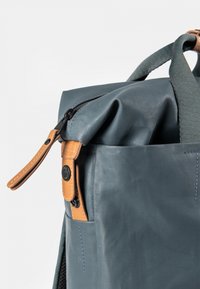 Rolltop-Rucksack aus dunkelblauem gummiertem Material mit Akzenten aus hellbraunem Leder, Reißverschluss, Seitentasche und strukturierter Oberfläche.