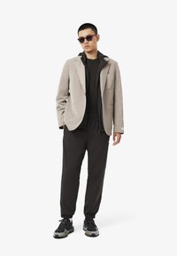 Lysegrå blazer over en sort lynlåsjakke, parret med sorte bukser med elastisk talje og sorte sneakers. Solbriller fuldender outfittet.