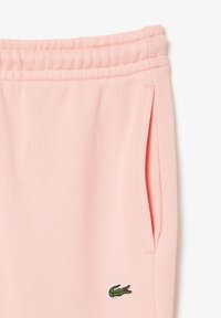 Pantalones de chándal de algodón rosa con cinturilla elástica, bolsillos laterales, textura acanalada y un logo de cocodrilo bordado en verde en la parte inferior de la pierna.