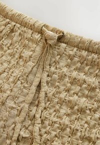 Strickstoff in Beige mit einem strukturierten Muster und gedrehten sowie verknoteten Details; elastischer Bund mit einer Krawatte.