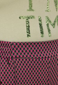 Korte mouwen top in lichtbeige met groene glitters tekst; gecombineerd met een patroonrok met geometrische vormen in roze en marineblauw.