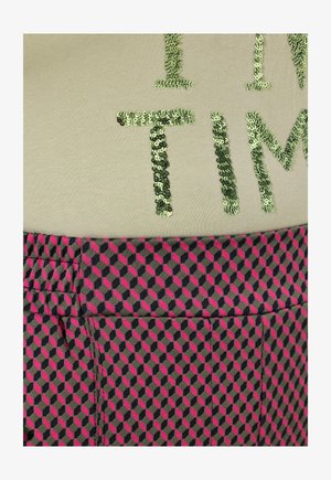 Kortærmet bluse i lys beige med grønne palietskrifter; parret med mønstret nederdel med geometriske former i pink og marineblå.