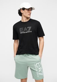Zwart katoenen t-shirt met witte "EA7 Emporio Armani"-logo; gecombineerd met muntgroene short met hetzelfde logo in het wit aan de zijkant.