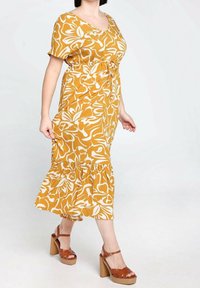 Żółta sukienka w kwiatowy wzór z dekoltem w kształcie litery V, krótkimi rękawami, elastycznym pasem i falbaną u dołu. Uzupełniona brązowymi sandałami na obcasie.