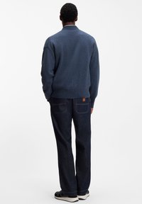 Blå tröja med hög krage och texturerad stickning; kombinerad med mörkblå jeans med två bakfickor. Skorna är svarta med vita sulor.