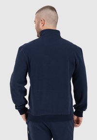Giacca in pile blu navy con colletto alto, maniche lunghe e polsini a costine. Presenta un'etichetta sul fondo a sinistra.
