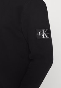 Zwarte lange mouw van een overhemd met een rechthoekige Calvin Klein Jeans logo patch in zwart, wit en grijs op de bovenarm.