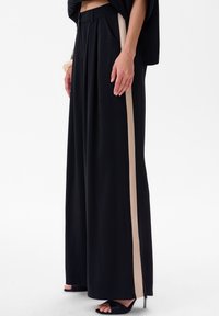 Zwarte wijde broek met een hoge tailleband, voorzien van beige zijstrepen, plooien en een zachte textuur. Gestyled met zwarte open-taille hakken.