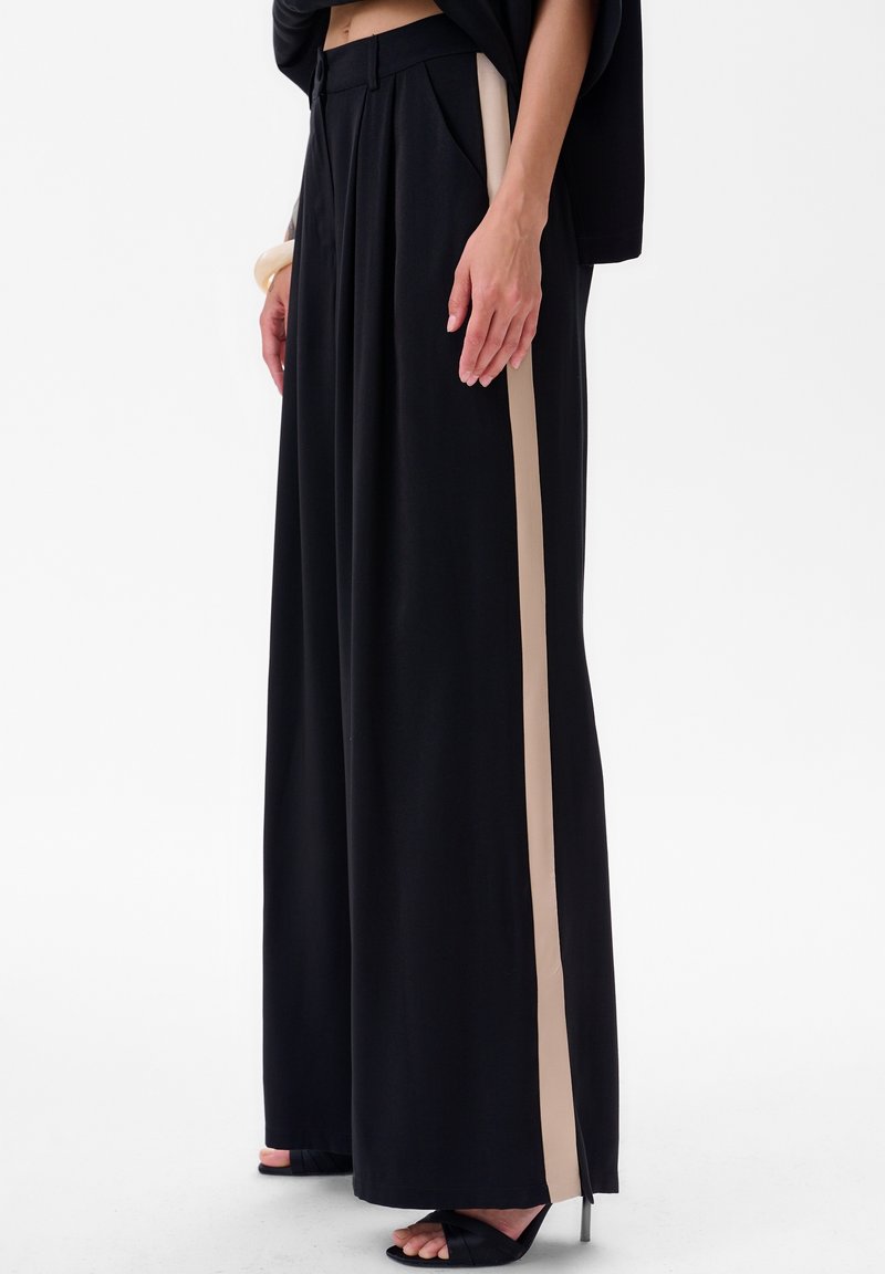 Zwarte wijde broek met een hoge tailleband, voorzien van beige zijstrepen, plooien en een zachte textuur. Gestyled met zwarte open-taille hakken.