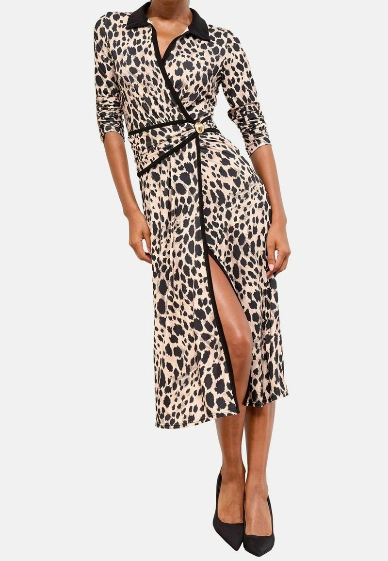 Femme portant une robe portefeuille à imprimé léopard avec un col noir, un bord noir, une boucle dorée et une fente sur le devant, assortie à des talons noirs pointus.
