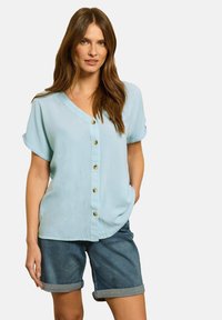 Blouse bleu clair à manches courtes, avec un col en V, des boutons à l'avant et des manches retroussées, associée à un short en denim foncé roulé au bord.