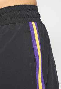 Bermudas atléticas pretas com um cós elástico, apresentando uma faixa lateral em roxo e amarelo, exibindo uma textura suave e um design moderno.