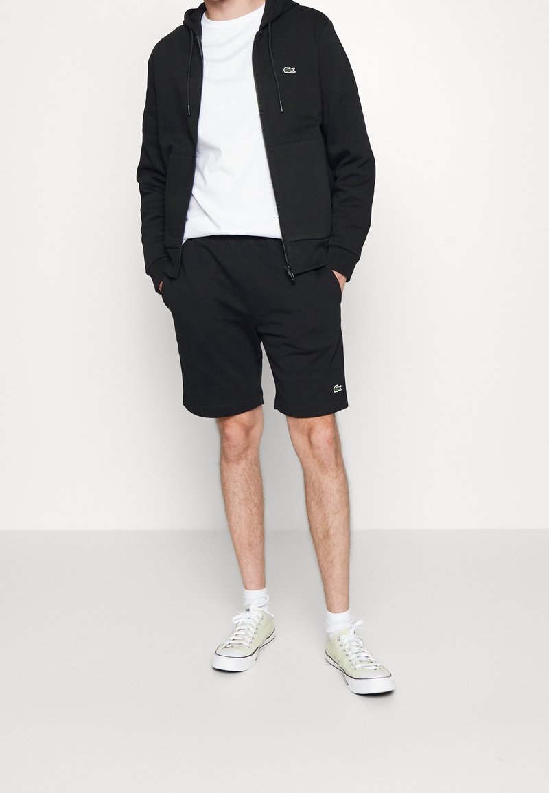 Svart zip-up hoodie och svarta shorts, båda gjorda av mjukt material. Vit t-shirt under. Sneakers i ljusgrön färg med vit sula.