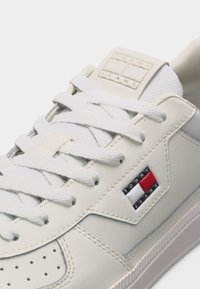 Λευκό αθλητικό παπούτσι Tommy Jeans με κορδόνια, ραμμένες λεπτομέρειες και σήμα λογότυπου στο πλάι και στη γλώσσα, κοντινή άποψη.